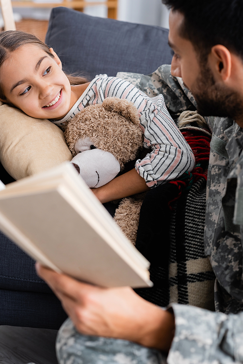 Happy child with teddy bear looking at blurred father in military uniform holding book at home