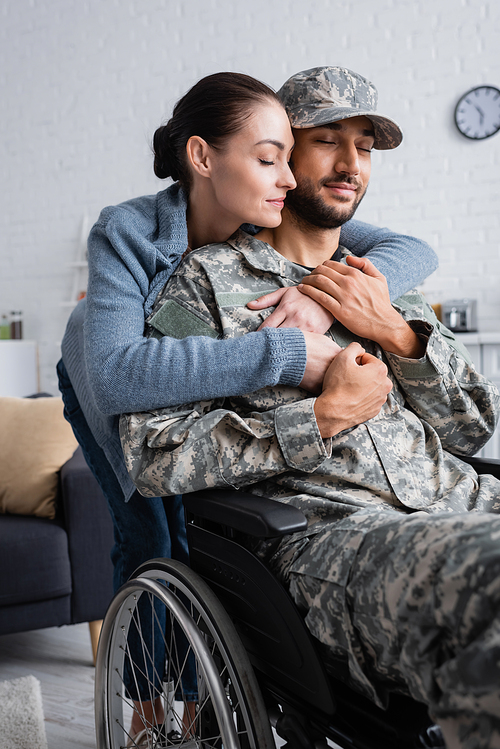 Woman hugging husband in camouflage uniform and wheelchair at home