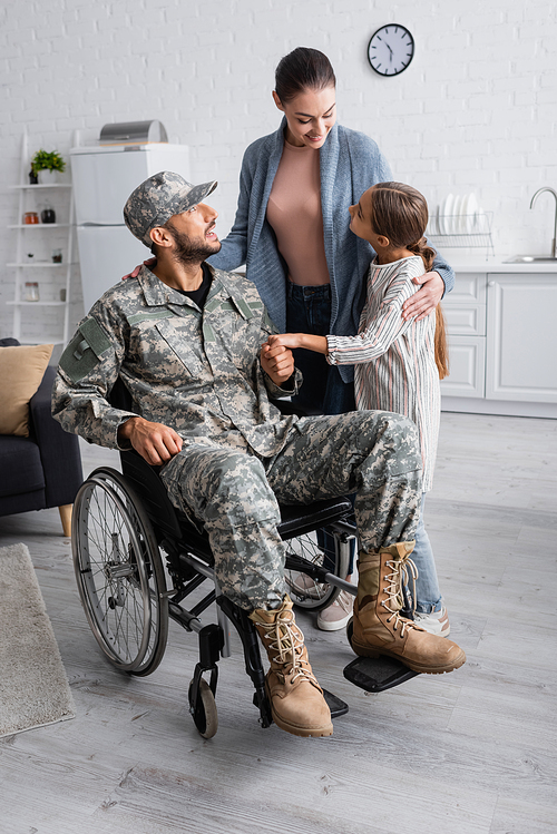Cheerful woman and child standing near father in military uniform and wheelchair at home