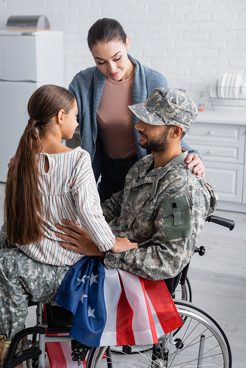 Woman and kid looking at father in military uniform and wheelchair with american flag at home