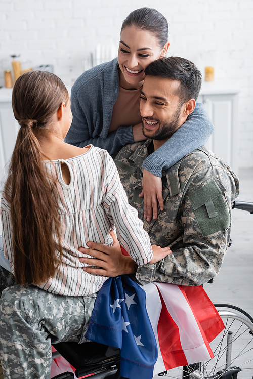 Smiling man in military uniform and wheelchair with american flag looking at kid near wife at home