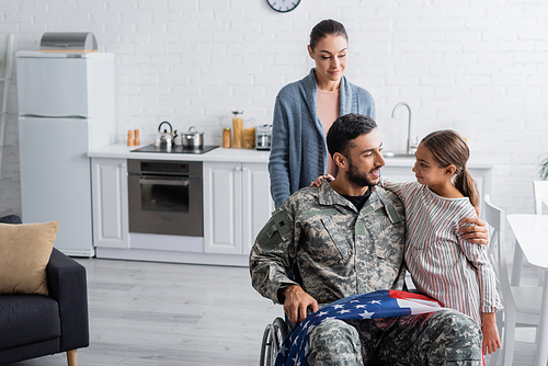 Smiling veteran in wheelchair with american flag hugging daughter near wife at home