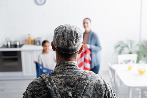 Soldier in camouflage standing near blurred family with american flag at home