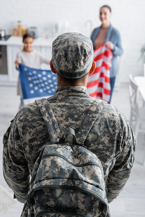 Man in camouflage uniform with backpack near blurred family with american flag at home