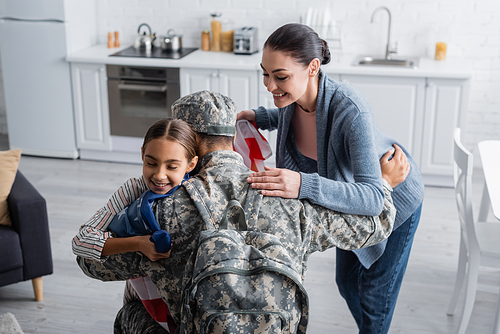 Smiling woman holding american flag while daughter hugging father in military uniform at home