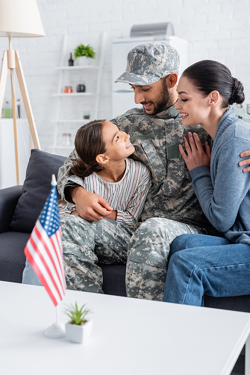 Positive man in military uniform hugging wife and kid near american flag at home