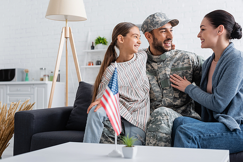Happy family hugging man in camouflage uniform on couch near american flag at home