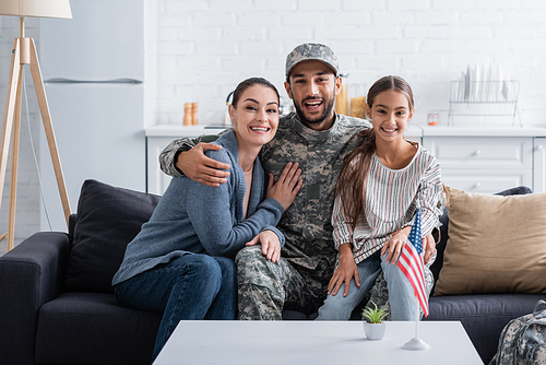 Positive man in camouflage uniform hugging family on couch near american flag at home