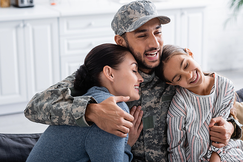 Joyful man in military uniform hugging daughter and wife at home