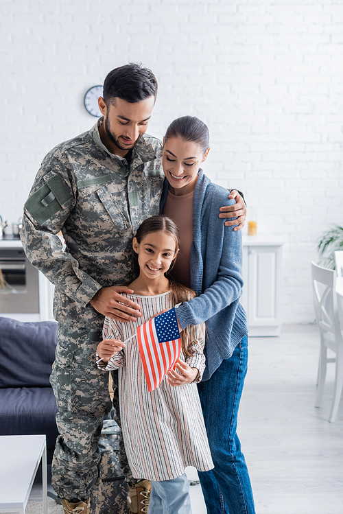 Smiling man in military uniform hugging wife and kid with american flag at home