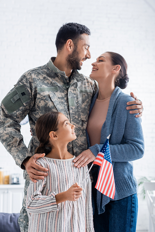 Positive woman looking at husband in camouflage uniform near daughter with american flag at home