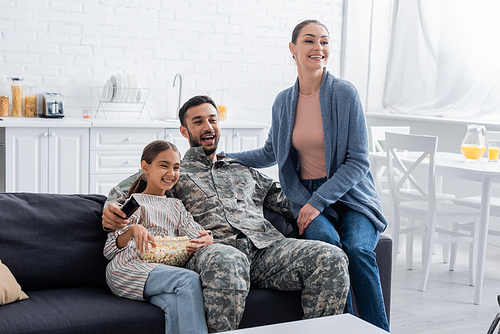 Smiling man in military uniform holding remote controller near family with popcorn at home