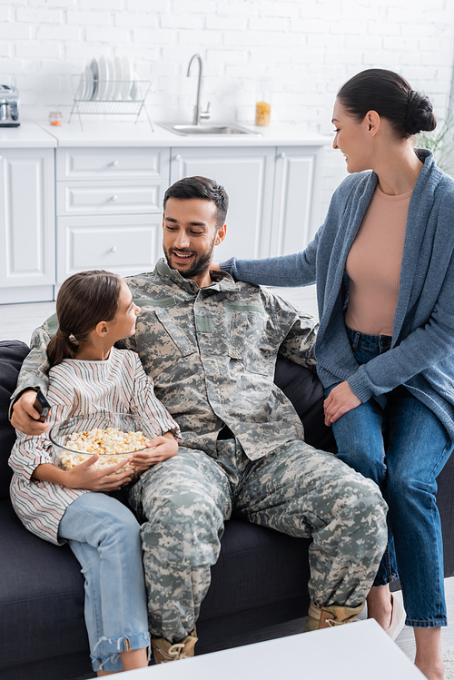 Soldier holding remote controller near daughter with popcorn and wife at home