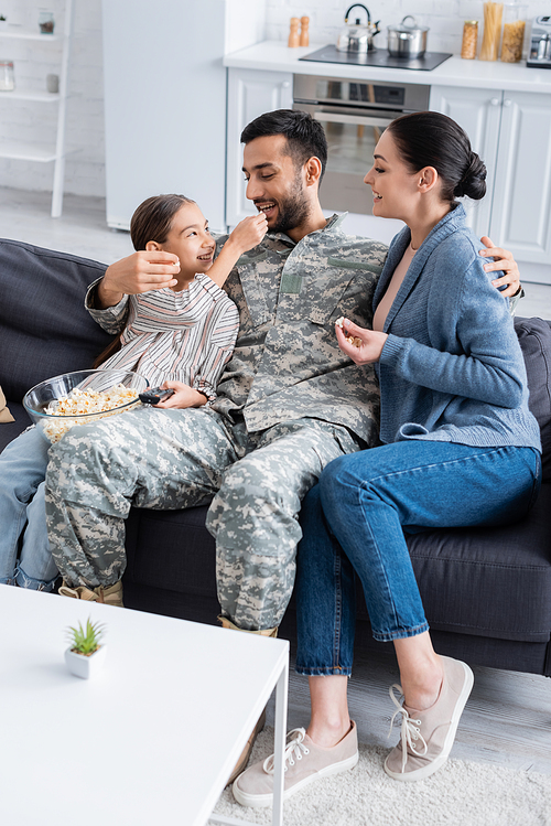 Kid holding popcorn and remote controller near father in military uniform and mom on couch