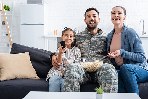 Smiling family with popcorn watching movie near father in camouflage uniform at home