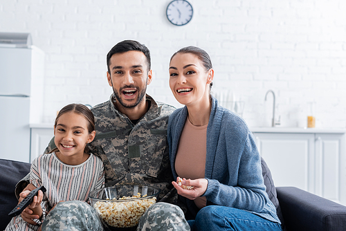 Positive man in military uniform sitting near family with remote controller and popcorn on couch