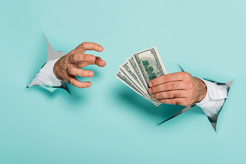 cropped view of man holding dollar banknotes through holes in paper wall on blue