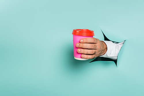 cropped view of man holding coffee to go through hole in paper wall on blue