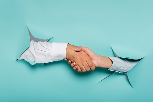 cropped view of man and woman shaking hands through holes in paper wall on blue