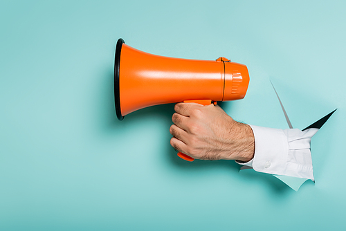 cropped view of man holding megaphone through hole in paper wall on blue