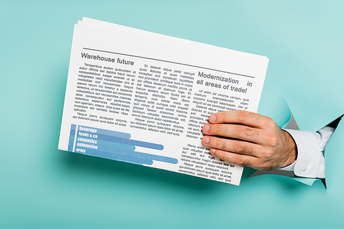 cropped view of man holding newspaper through hole in paper wall on blue