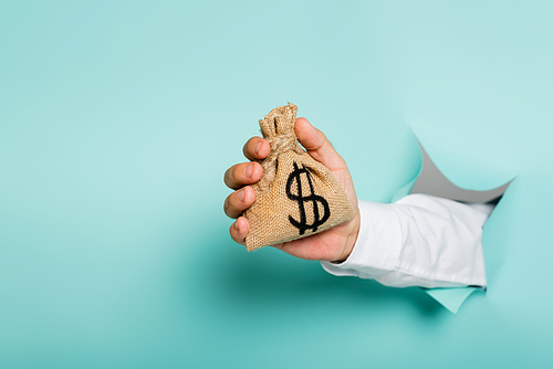 cropped view of man holding money bag with dollar sign through hole in paper wall on blue