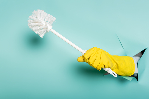 partial view of cleaner in rubber glove holding toilet brush through hole in paper wall on blue