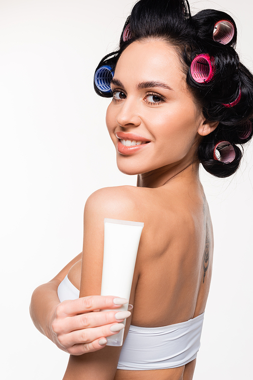 cheerful young woman in curlers and top holding cream tube near shoulder isolated on white