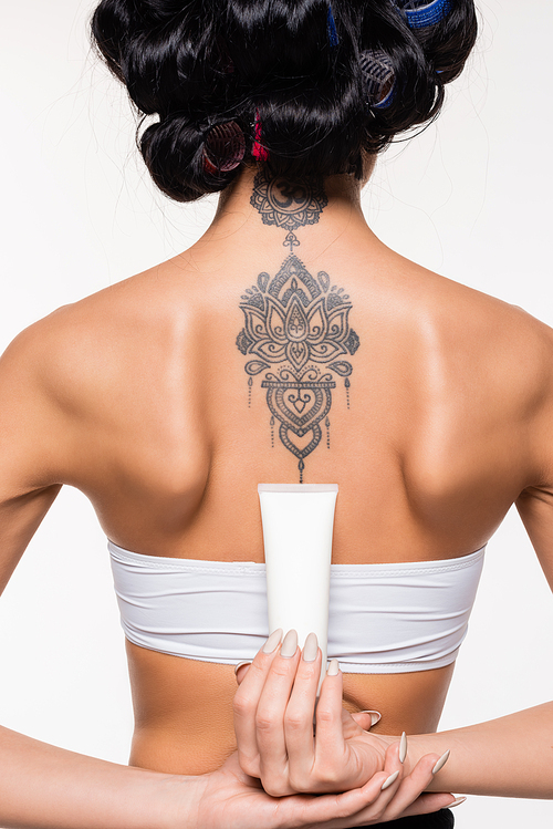 back view of young woman in curlers with tattoo holding cream tube isolated on white