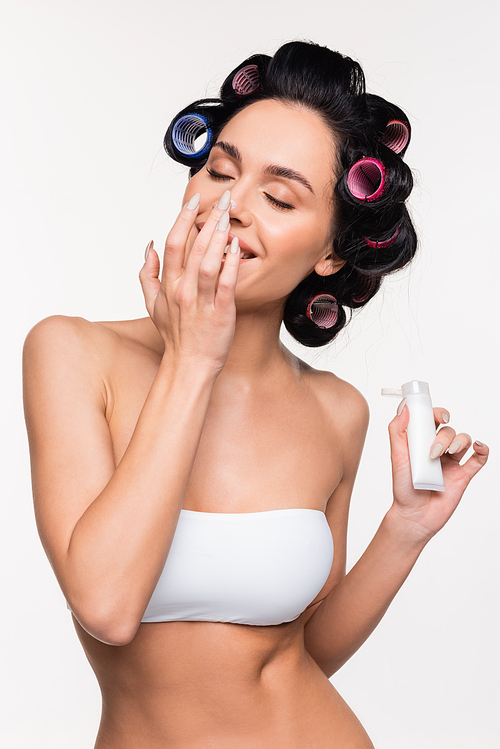 young woman with closed eyes in curlers and top applying cream on nose and holding tube isolated on white