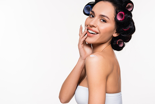 smiling young woman in curlers and top posing with applied cream on shoulder and hand near face isolated on white