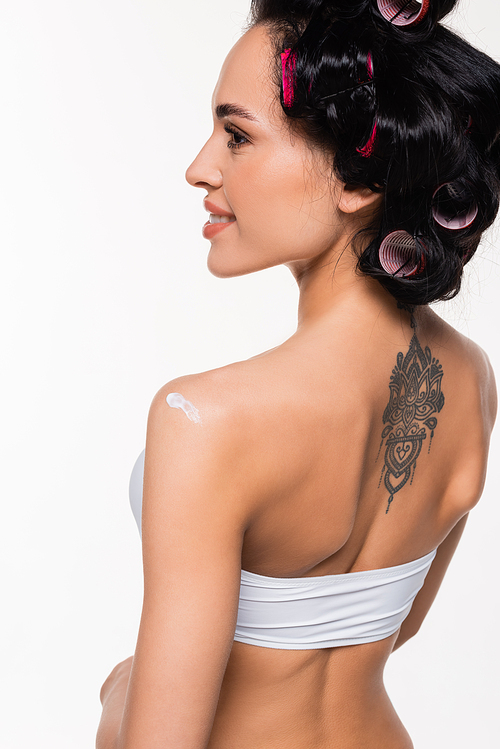 smiling young woman in curlers and tattoo on back standing with applied cream on shoulder isolated on white