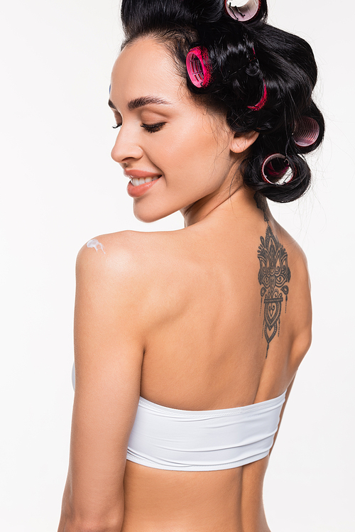 smiling young woman in curlers and tattoo on back standing with applied cream on shoulder isolated on white