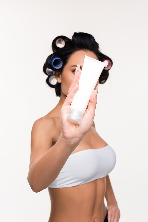 young woman in curlers covering face with cream tube in hand isolated on white