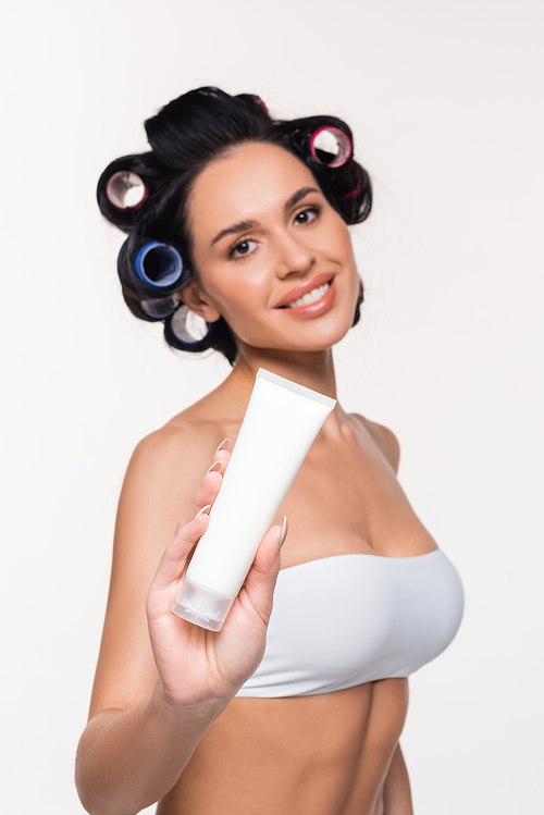 smiling young woman in curlers and bra showing cream tube isolated on white