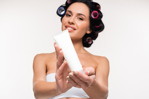 cheerful young woman in curlers and top showing cream tube isolated on white