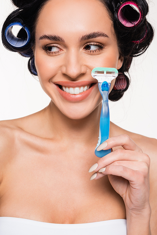 positive young woman in curlers holding razor near face isolated on white