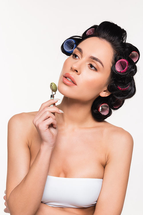 young woman in curlers making facial massage with jade roller isolated on white