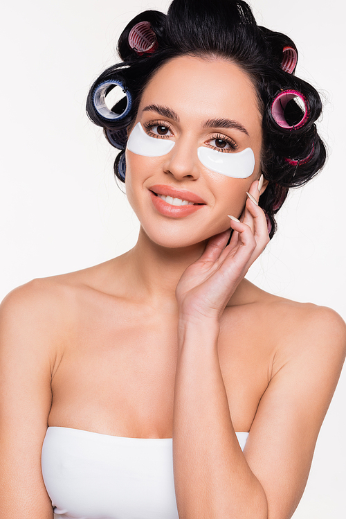 smiling young woman in curlers with eye patches holding hand near face isolated on white