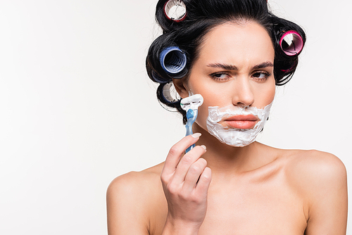 embarrassed young woman in curlers holding razor near face with shaving cream isolated on white