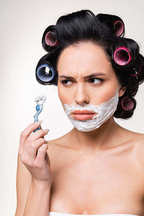 confused young woman in curlers holding razor near face with shaving cream isolated on white
