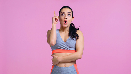 surprised young woman in sportswear pointing with finger and looking up isolated on pink