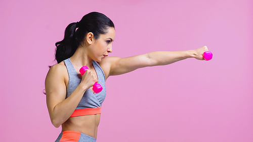 side view of strong young sportswoman working out with dumbbells isolated on pink