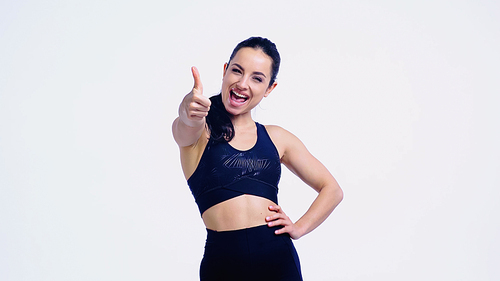 smiling sportswoman showing thumb up while standing with hand on hip isolated on white