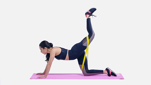 brunette young sportswoman working out with yellow elastics on pink fitness mat on white