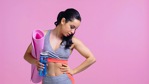 tired sportswoman holding sports bottle and fitness mat while standing with hand on hip isolated on pink