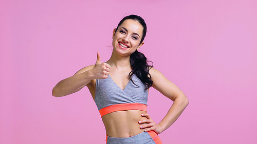 positive young sportswoman standing with hand on hip and showing thumb up isolated on pink