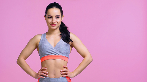 smiling young sportswoman standing with hands on hips isolated on pink