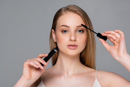 young woman holding tube and brush while applying mascara isolated on grey
