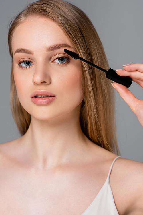 young woman holding brush and applying mascara isolated on grey
